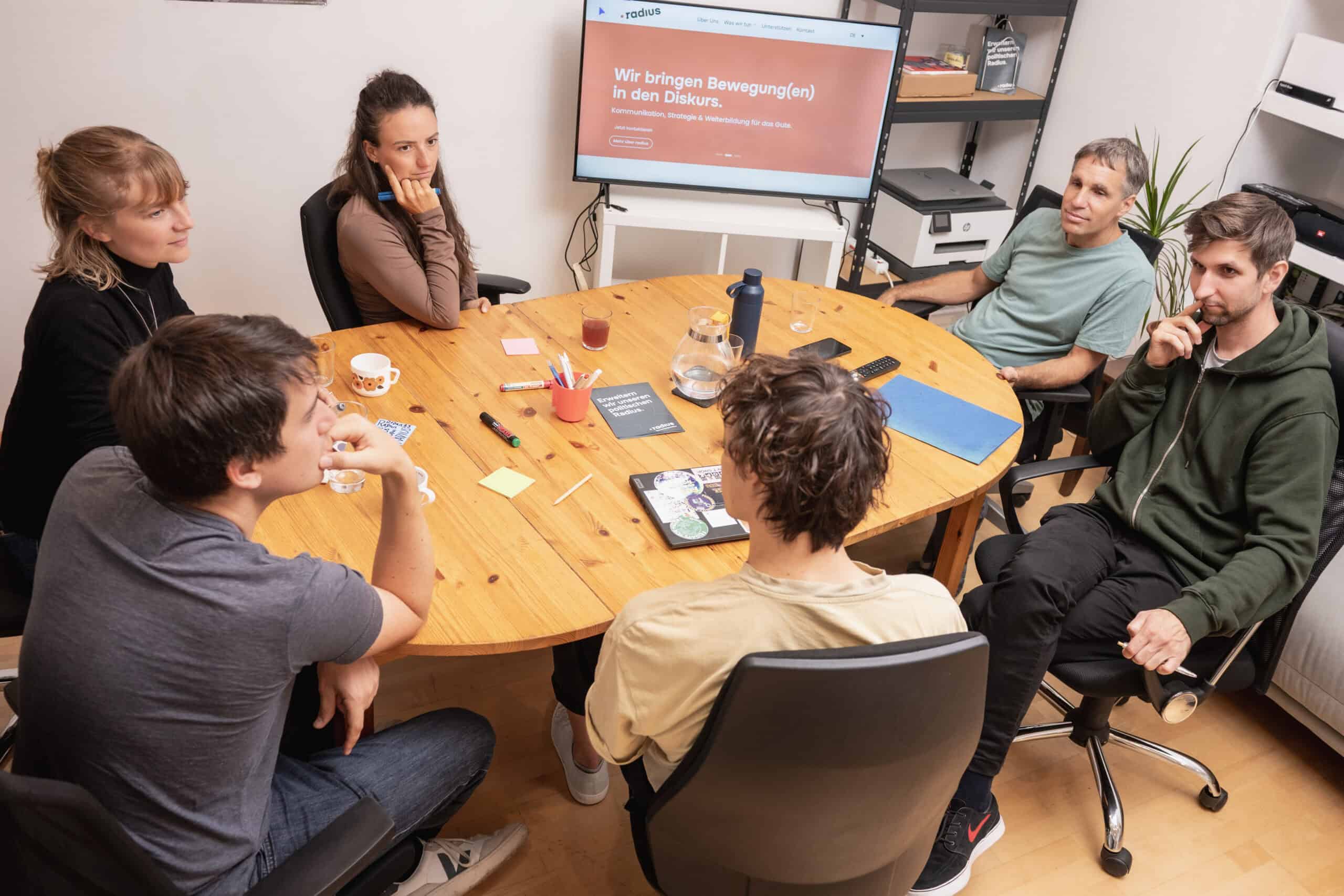 Besprechung im radius Team, ganzes Team ist um einen Tisch zu sehen, 6 Personen insgesamt um eine ovalen Tisch herum